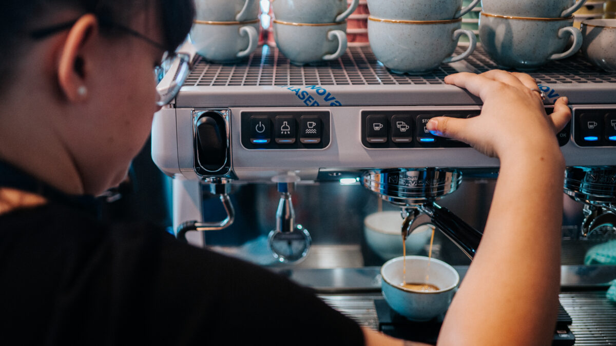 Od czego zależy cena kawy? Barista obsługujący ekspres do kawy w kawiarni, przygotowujący filiżankę kawy. W tle widoczne są różnokolorowe filiżanki ustawione na półce.