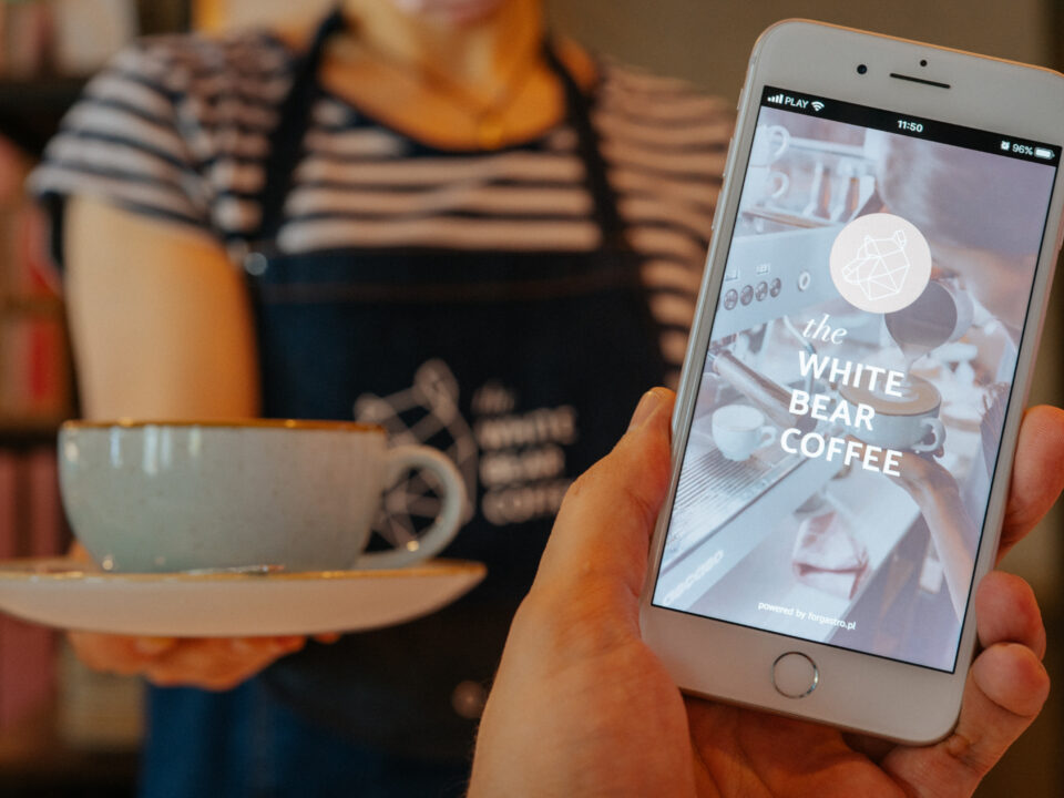 Kawa z dostawą Białystok. Sieć the White Bear Coffee weszła na rynek delivery. Osoba trzymająca filiżankę kawy na spodku, a w tle zbliżenie na ekran telefonu z logo i nazwą kawiarni 'The White Bear Coffee