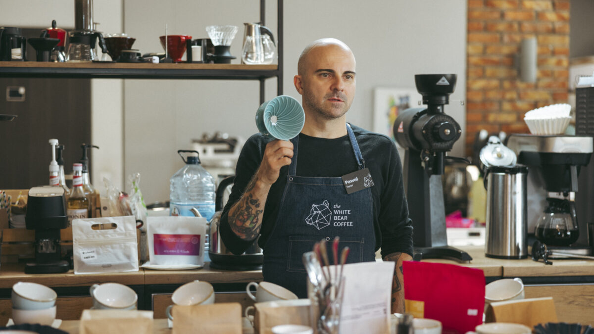 Nowy szkoleniowiec sieci The White Bear Coffee podczas warsztatów kawowych, trzymający w ręku ceramiczny dripper do parzenia kawy. Mężczyzna w fartuchu z logo firmy otoczony jest profesjonalnym sprzętem do parzenia kawy oraz różnymi akcesoriami baristycznymi na blacie