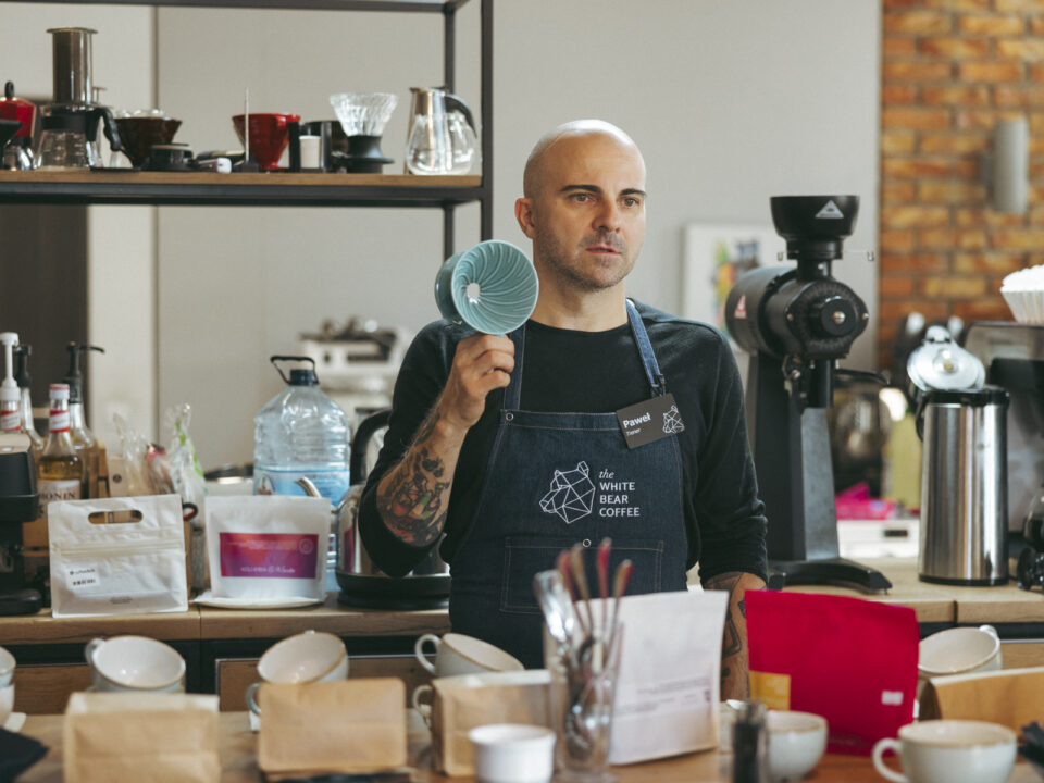 Nowy szkoleniowiec sieci The White Bear Coffee podczas warsztatów kawowych, trzymający w ręku ceramiczny dripper do parzenia kawy. Mężczyzna w fartuchu z logo firmy otoczony jest profesjonalnym sprzętem do parzenia kawy oraz różnymi akcesoriami baristycznymi na blacie