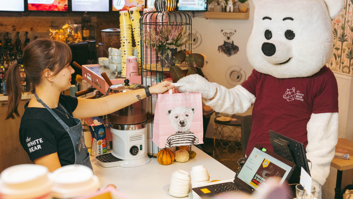 Baristka sieci the White Bear Coffee podająca człowiekowi w przebraniu misia polarnego torbę z kawą na wynos.