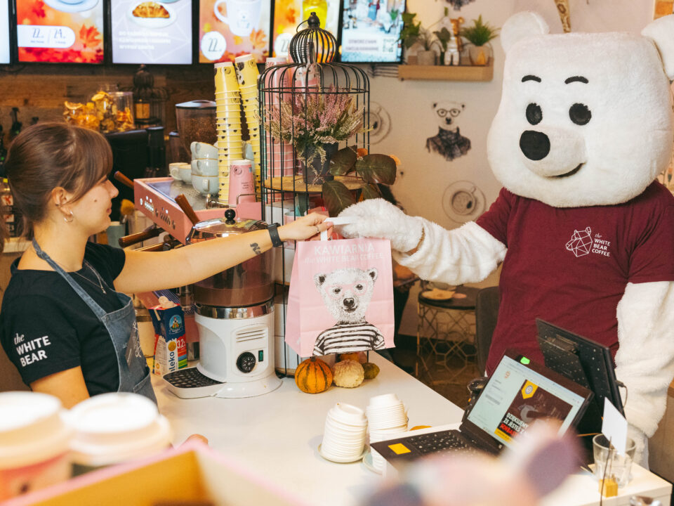 Baristka sieci the White Bear Coffee podająca człowiekowi w przebraniu misia polarnego torbę z kawą na wynos.